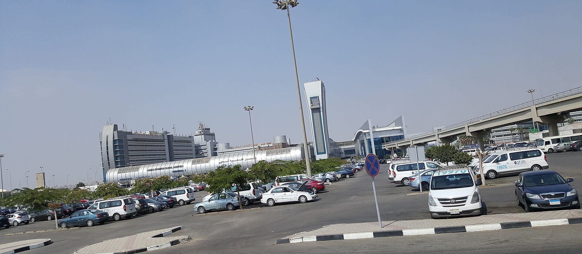The curved silver facade of Terminal 1 at Cairo International Airport seen across the car park, with taxis and minivans parked in front
