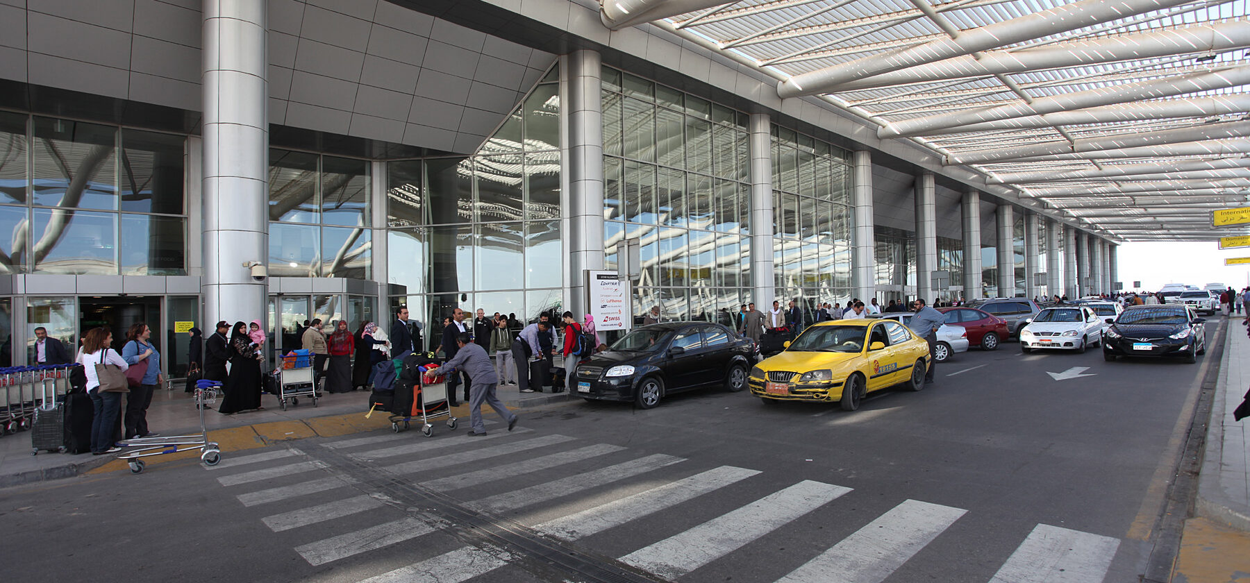 The kerbside of Terminal 3 at Cairo International Airport, with passengers, luggage trolleys and a yellow taxi under the steel canopy
