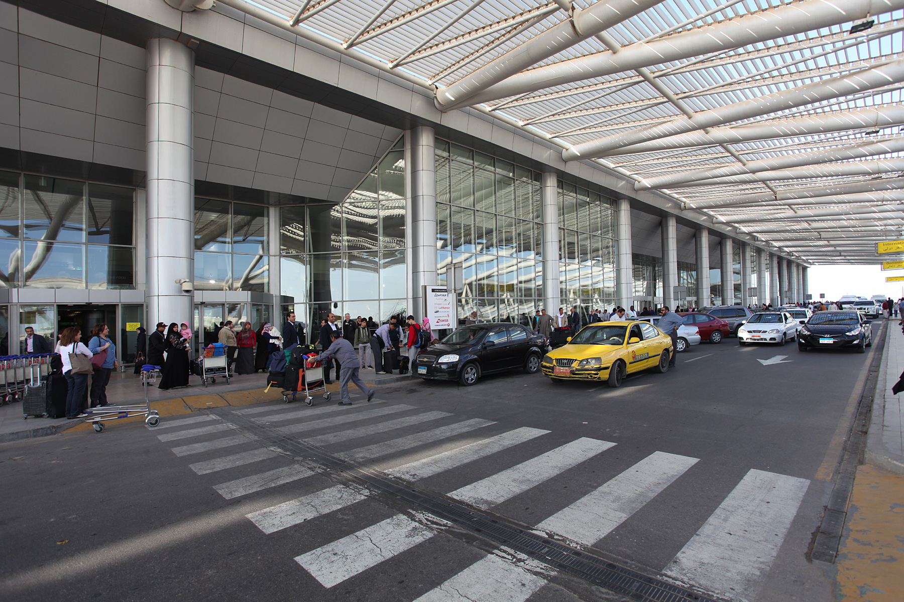 The kerbside of Terminal 3 at Cairo International Airport, with passengers, luggage trolleys and a yellow taxi under the steel canopy