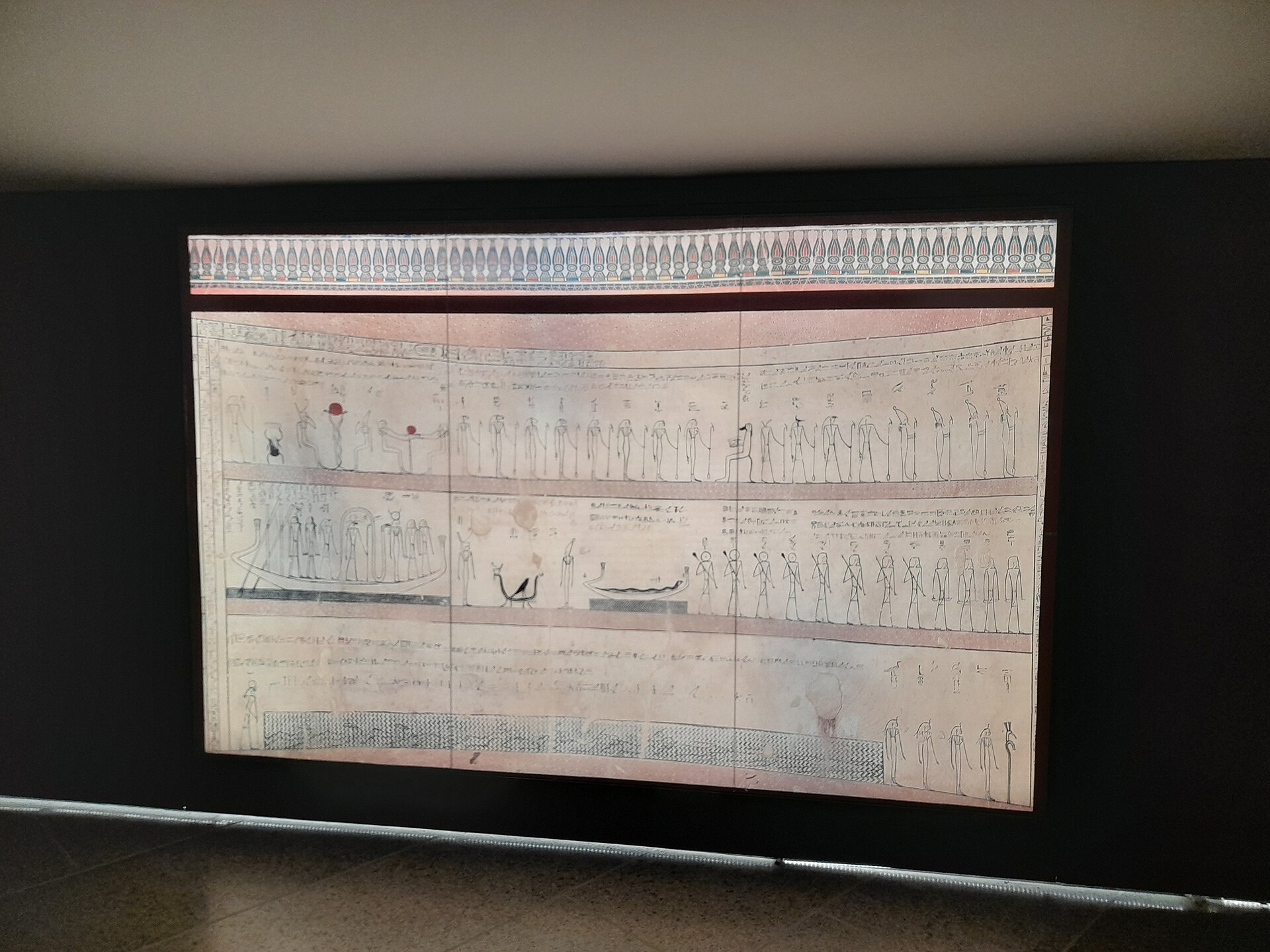 Illuminated display of a painted funerary papyrus with rows of deities in a darkened gallery at the Grand Egyptian Museum