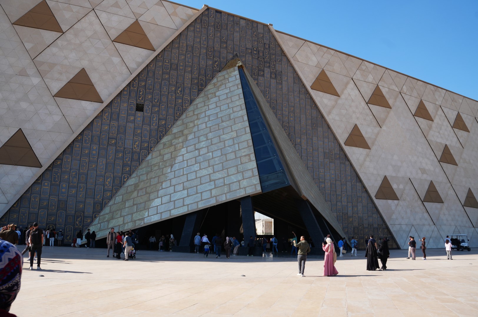 Grand Egyptian Museum Tickets and Prices: How to Book Online 2 Visitors walking towards the triangular main entrance of the Grand Egyptian Museum below its hieroglyph-carved stone facade