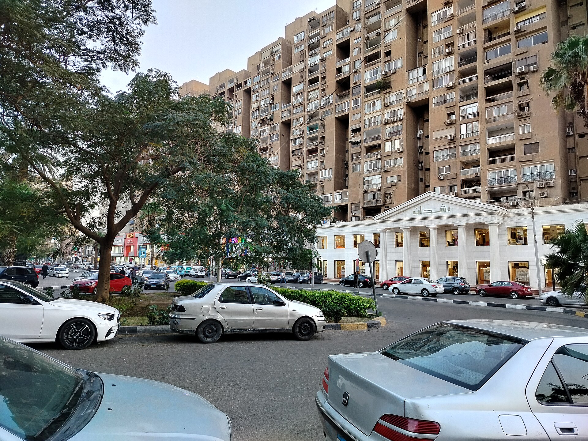 A tree-lined street in Heliopolis, Cairo, with tall apartment blocks and a white colonnaded shopfront