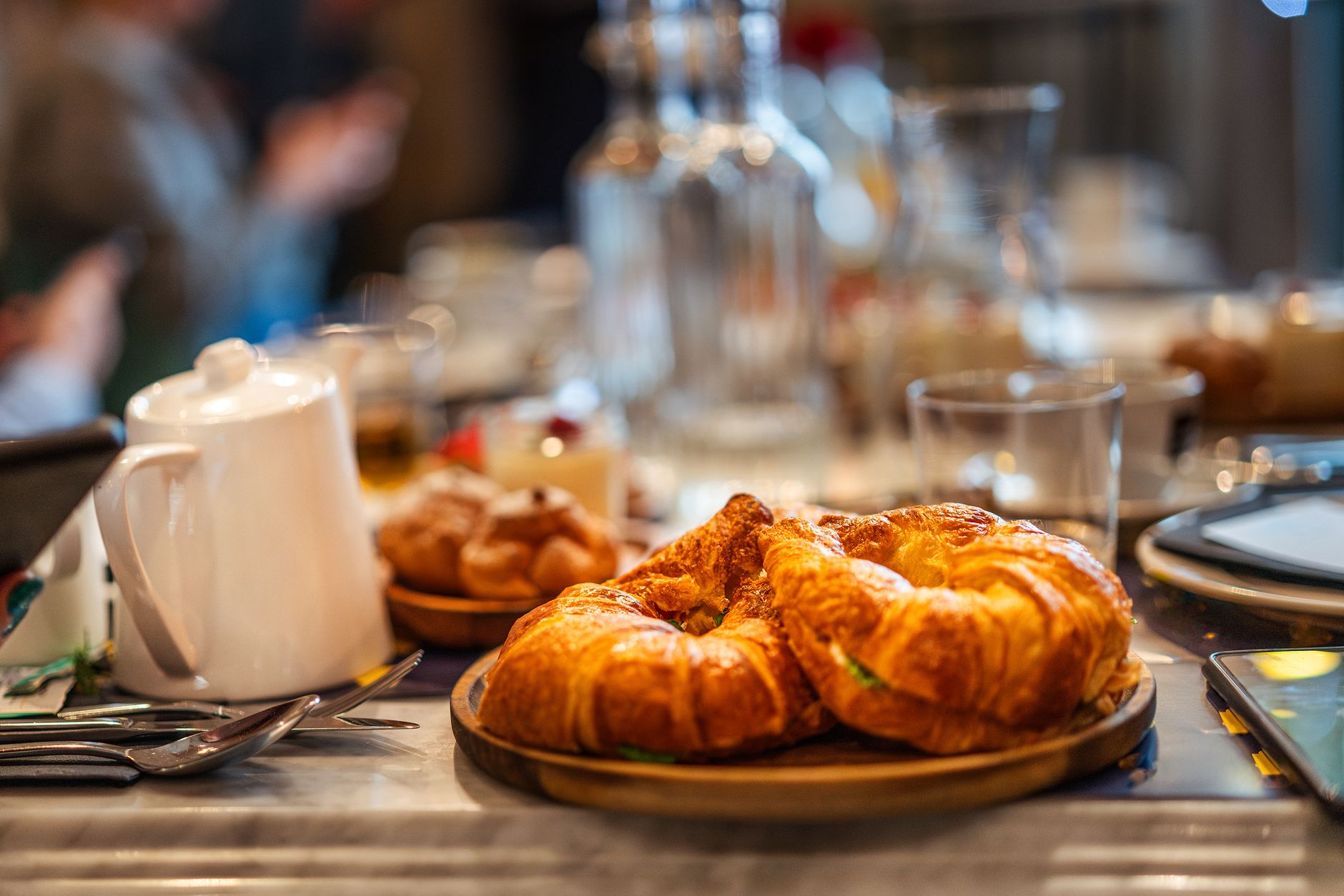 Croissants on a wooden board beside a white teapot and glasses on a café table