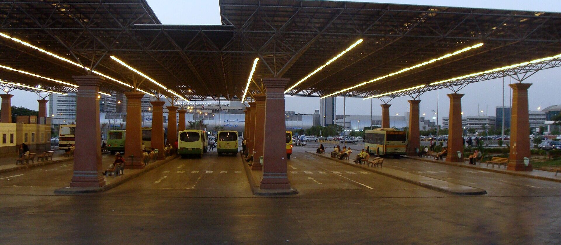 The covered bus station at Cairo International Airport, with buses parked beneath a long steel canopy on brick pillars at dusk