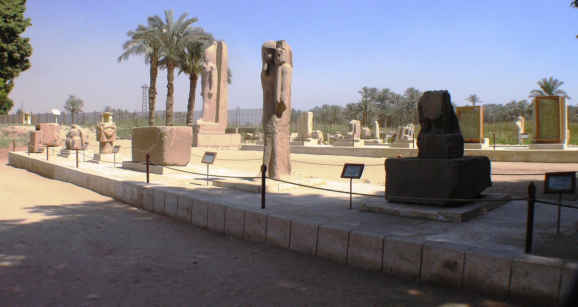 Statues, stelae and sculpture fragments displayed along a paved path among palm trees in the open-air museum at Memphis