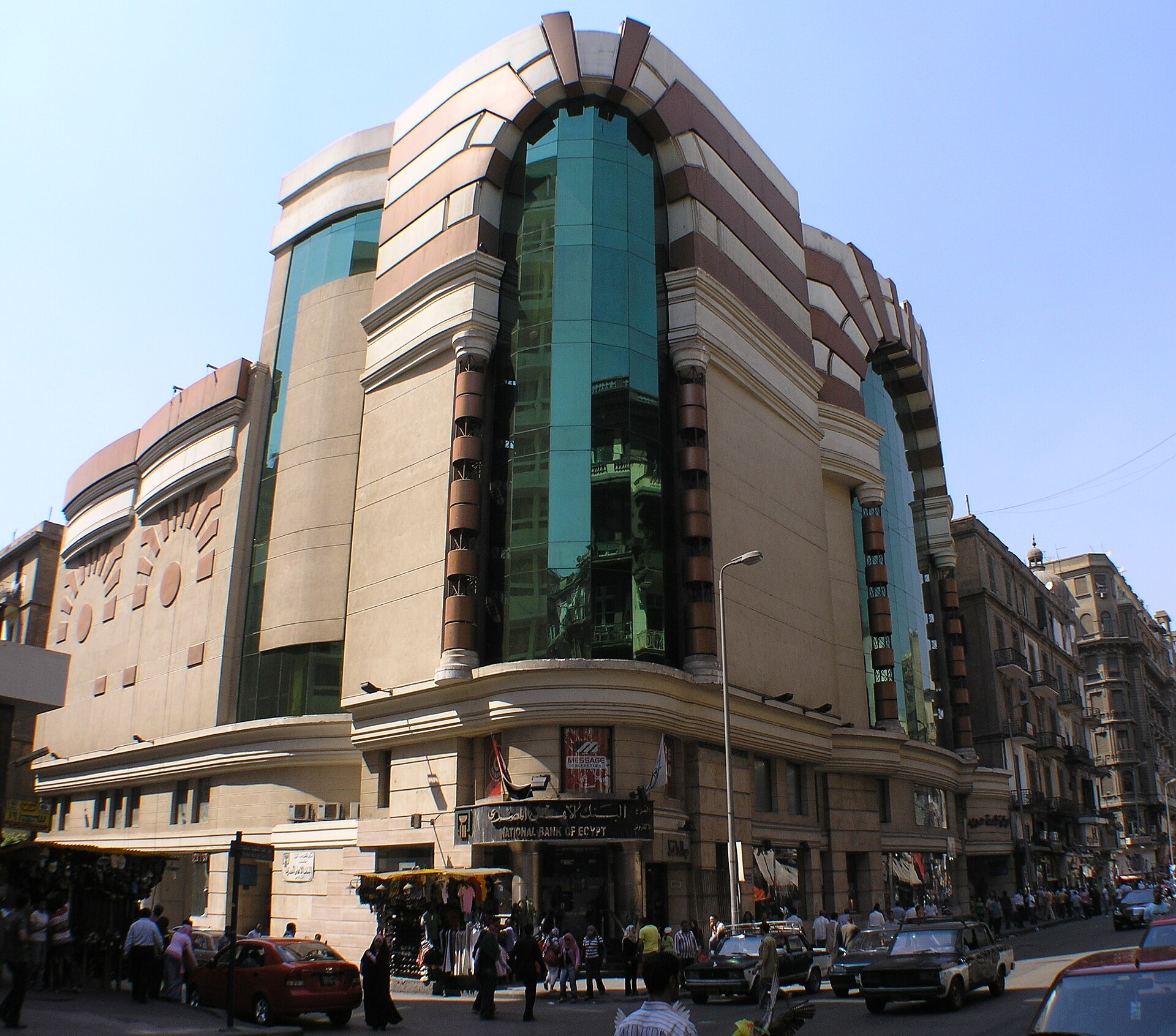A National Bank of Egypt branch at street level beneath the curved glass and stone facade of Talaat Harb Mall in Downtown Cairo, with shoppers and cars on the street